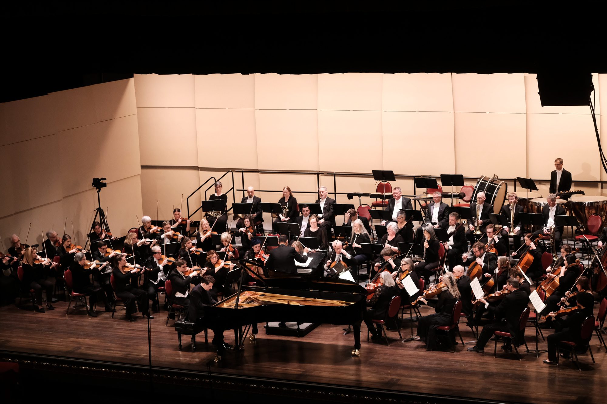 A pianist performs with the Saginaw Bay Symphony Orchestra, with the conductor and musicians visible across the stage.